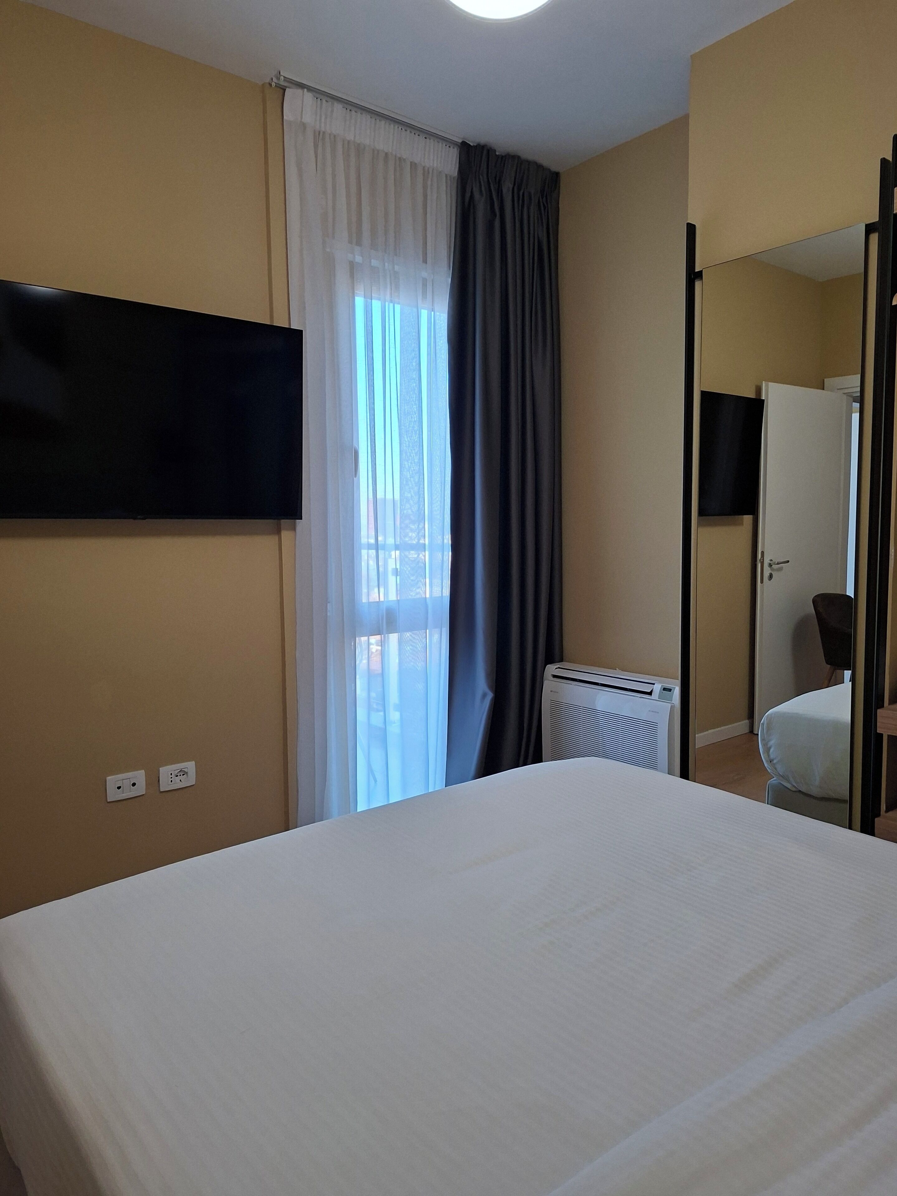 A hotel room with a bed, a television mounted on the wall, a window with curtains, a radiator, and a door.