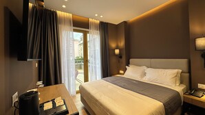 Habitación de hotel con una cama grande, un escritorio con cafetera, televisor montado en la pared y un balcón con cortinas.