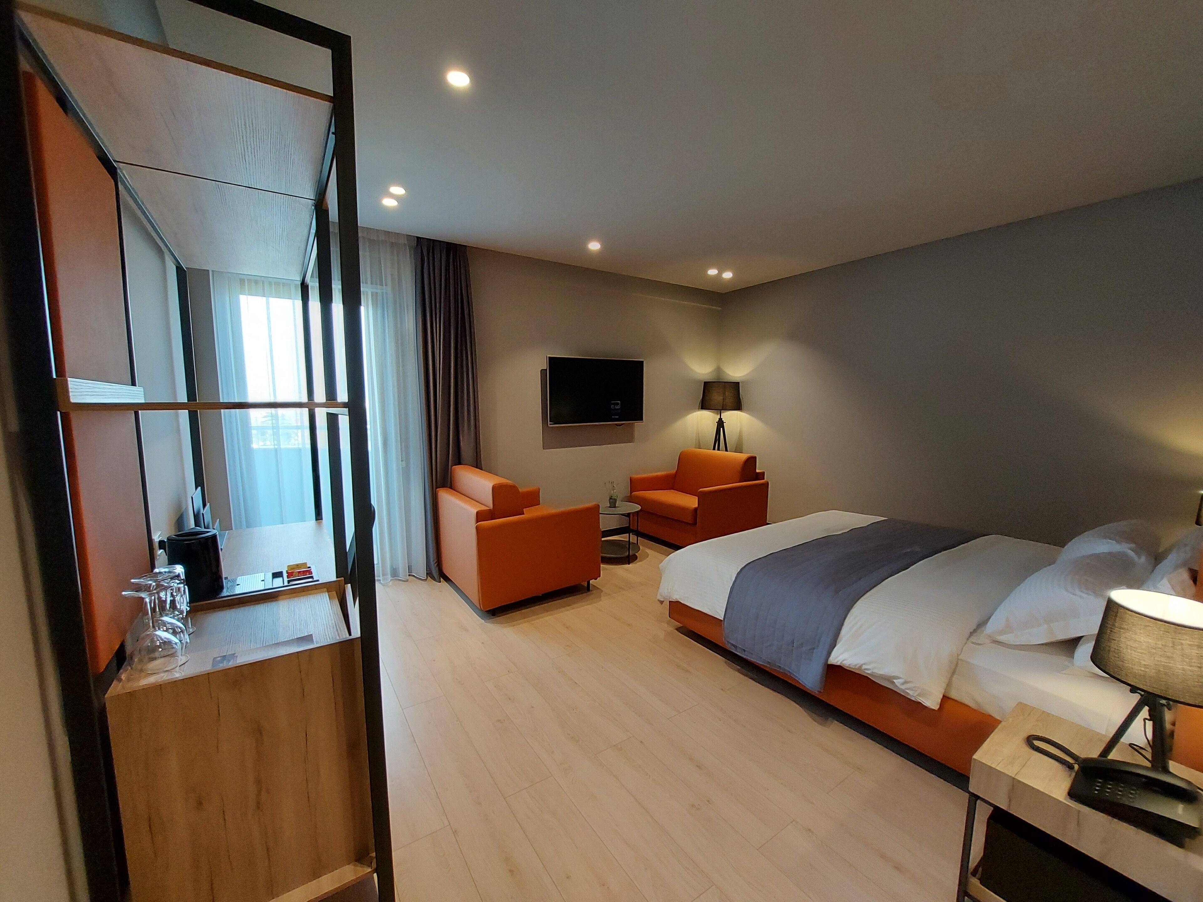 A hotel room with a bed, two orange armchairs, a small table, a lamp, a TV, and a mirror.