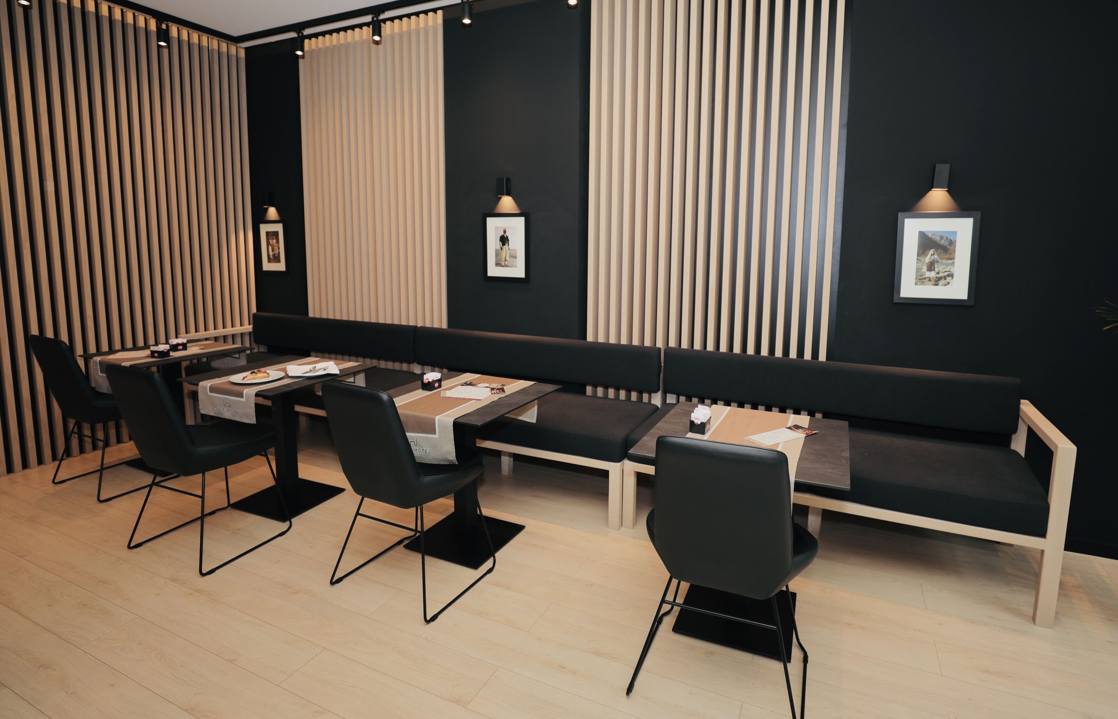A modern dining area with a long table, black chairs, and a bench. There are framed pictures on the wall and a pendant light above the table.