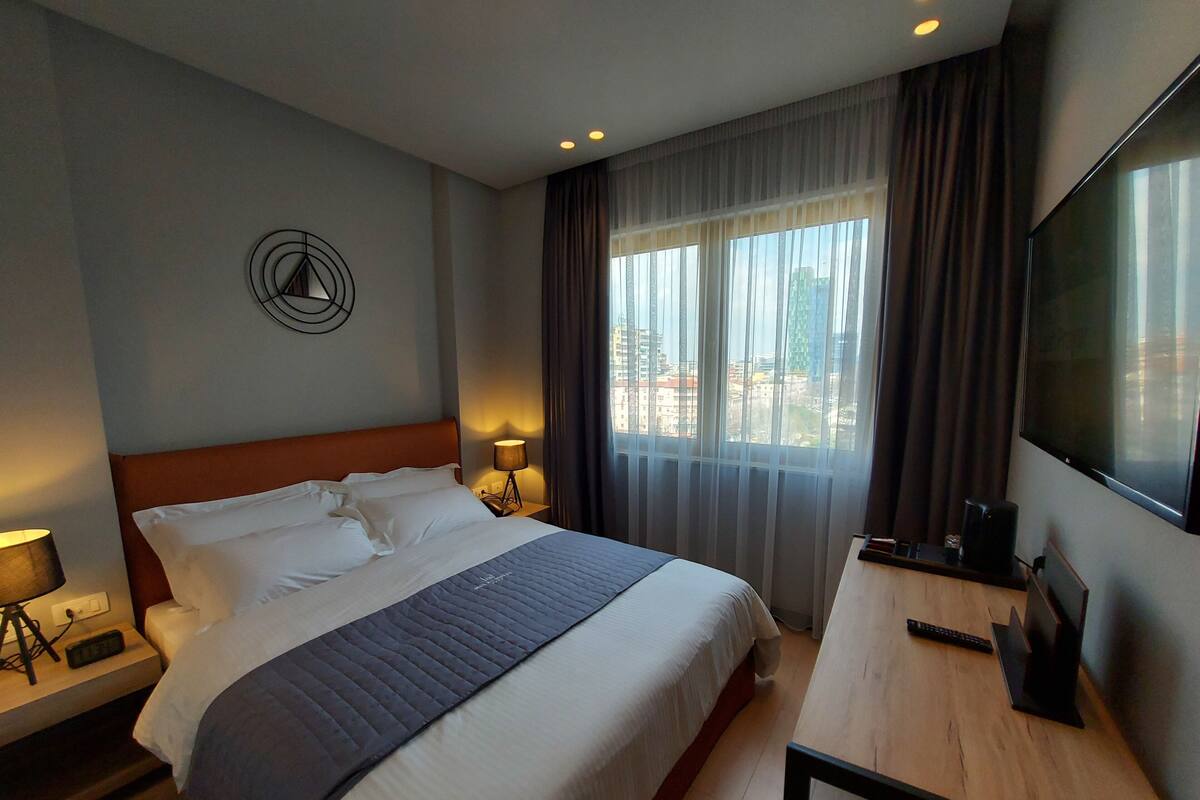 A hotel room with a large bed, a desk with a TV, and a view of buildings through the window.