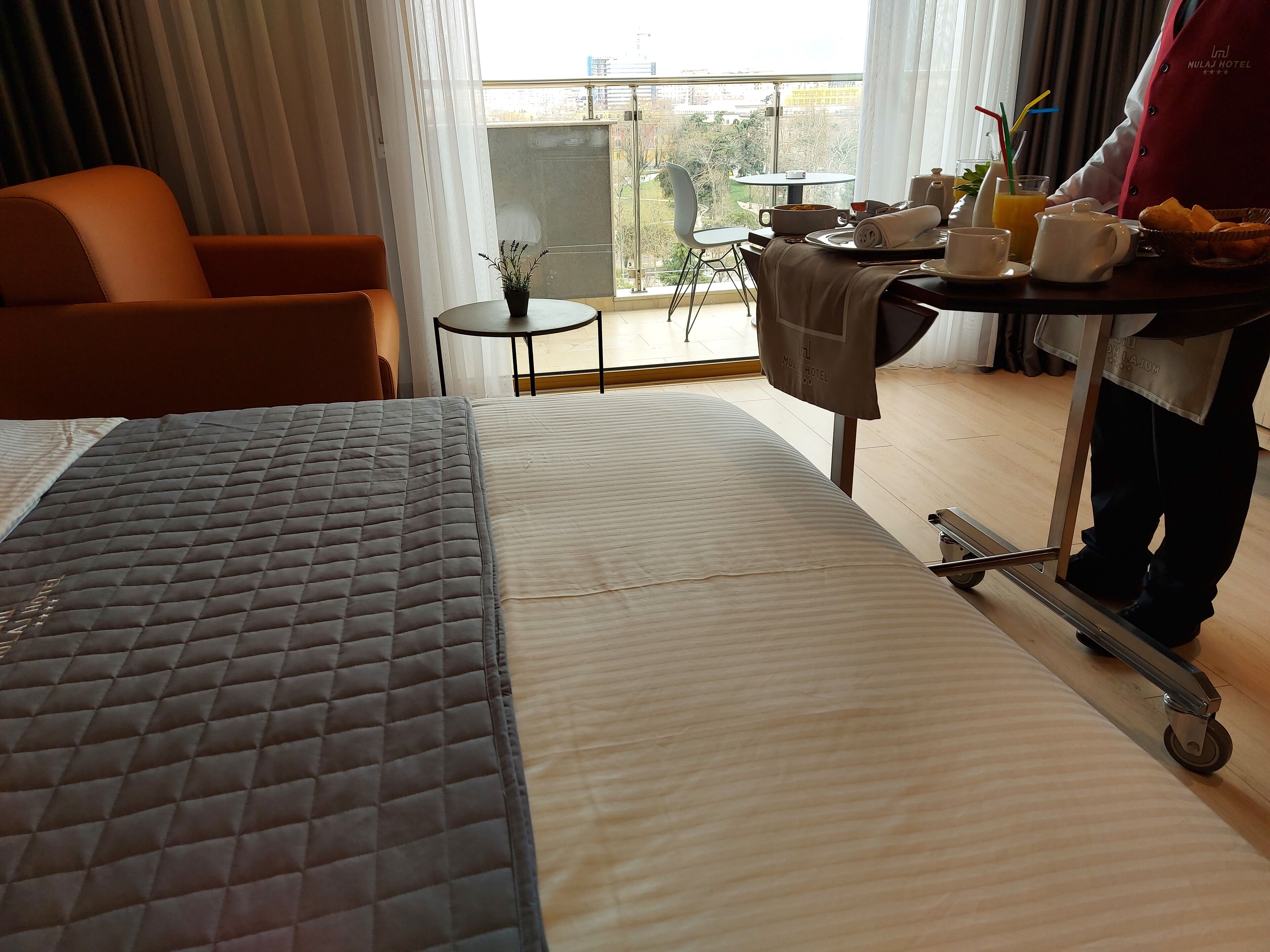 A hotel room with a bed, a chair, a small table with breakfast items, and a view of the city through large windows.
