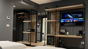 Una habitación de hotel moderna con dos camas, un televisor montado en un estante de madera, una caja fuerte y un mueble pequeño con vasos y una cafetera.