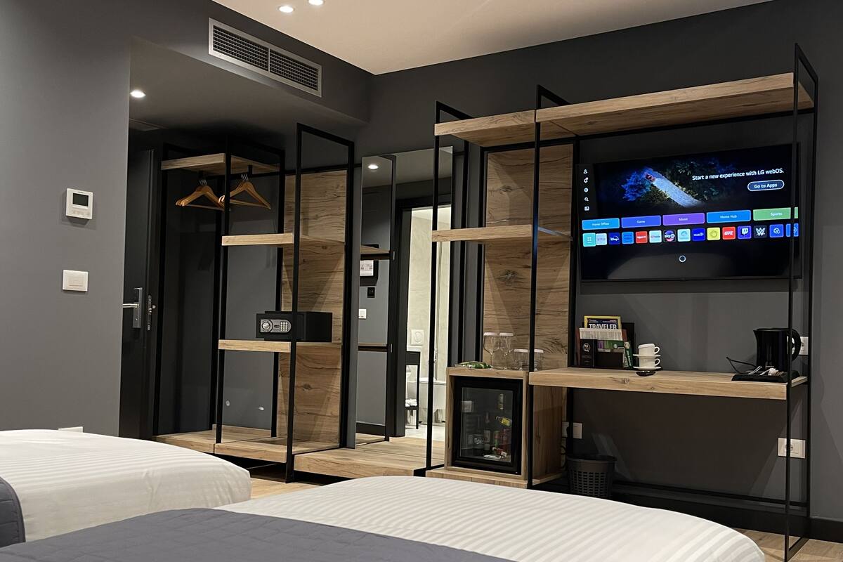 A modern hotel room with two beds, a TV mounted on a wooden shelf, a safe, and a small cabinet with glasses and a coffee maker.