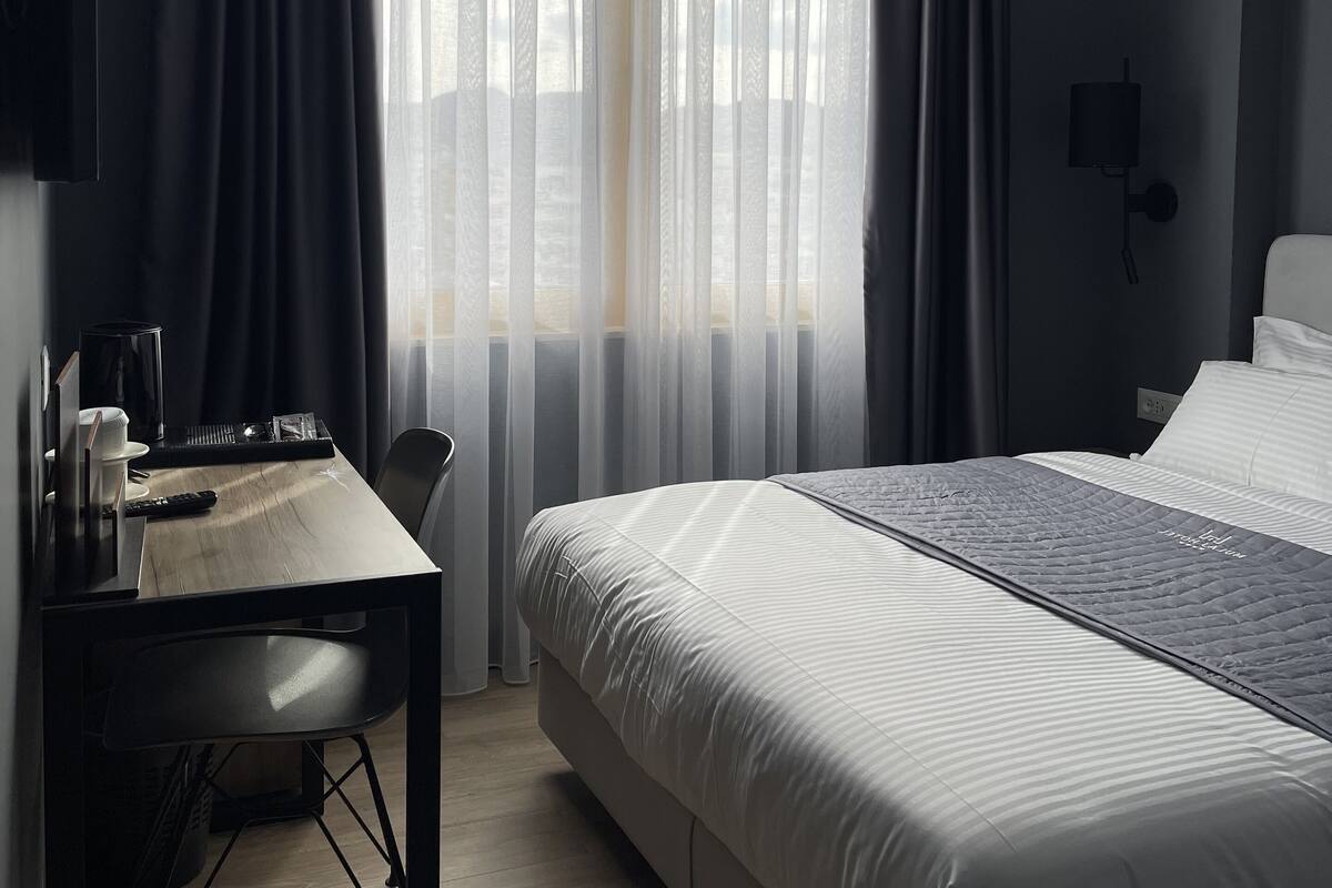 A modern hotel room with a large bed, a desk with a chair, a television, and a window with sheer curtains.