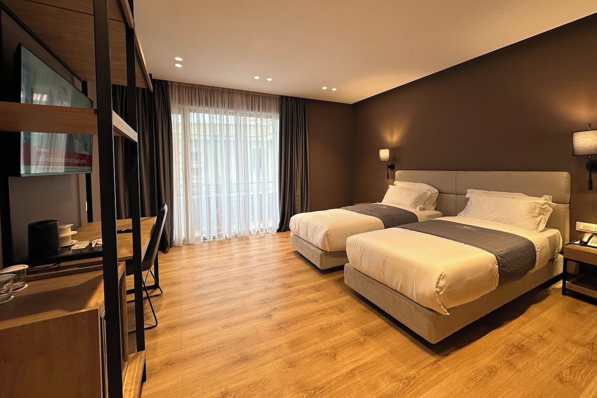 A hotel room with two beds, a wooden floor, a large window with curtains, and a flat-screen TV on a wooden shelf.