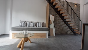 A modern living room with a glass-top coffee table, a grey sofa with striped cushions, and a wooden staircase against a stone wall. - Alios Gaia (Sithonia)