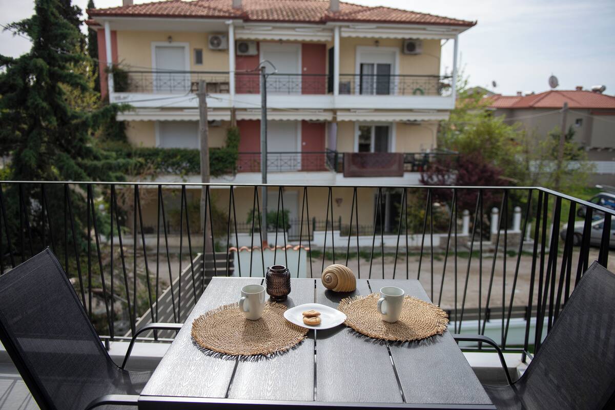 A balcony with a table set for two, featuring two cups, a plate with cookies, and a decorative shell. The balcony overlooks a building with multiple balconies and a tiled roof.