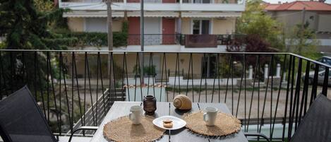 Un balcon avec une table dressée pour deux personnes, agrémentée de deux tasses, d’une assiette de biscuits et d’un coquillage décoratif. Le balcon offre une vue sur un immeuble doté de nombreux balcons et d’un toit recouvert de tuiles.