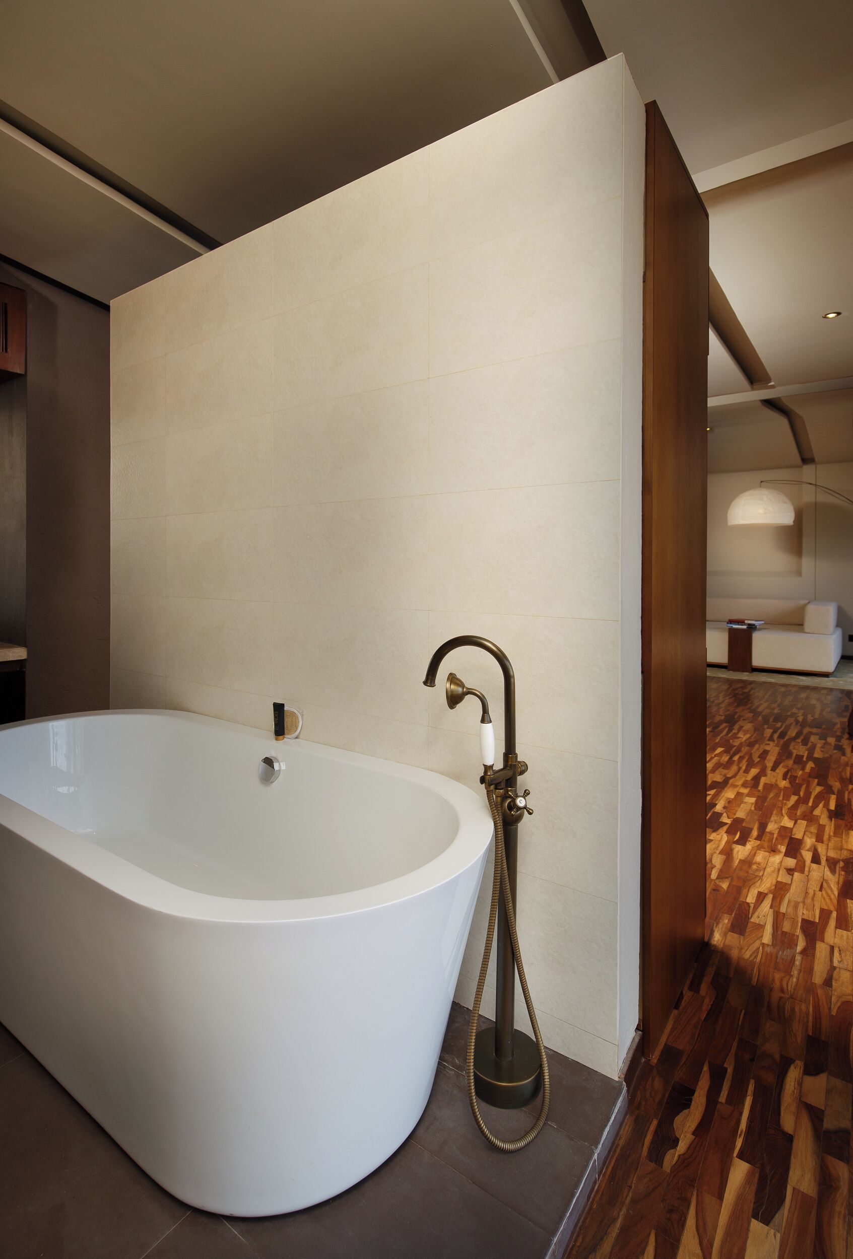 A modern bathroom with a freestanding bathtub, a wooden floor, and a visible living area with a sofa and lamp.