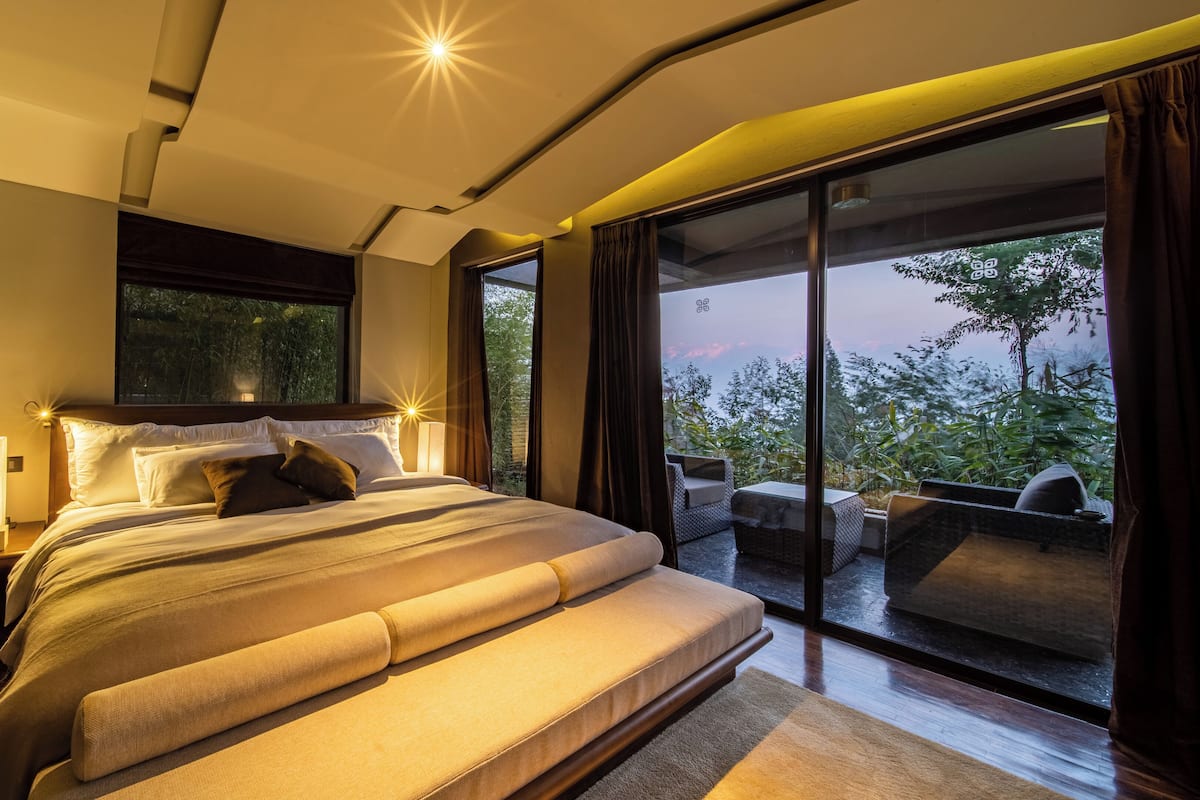 A modern bedroom with a large bed, a nightstand, a bedside lamp, and a view of the outdoors through a sliding glass door.