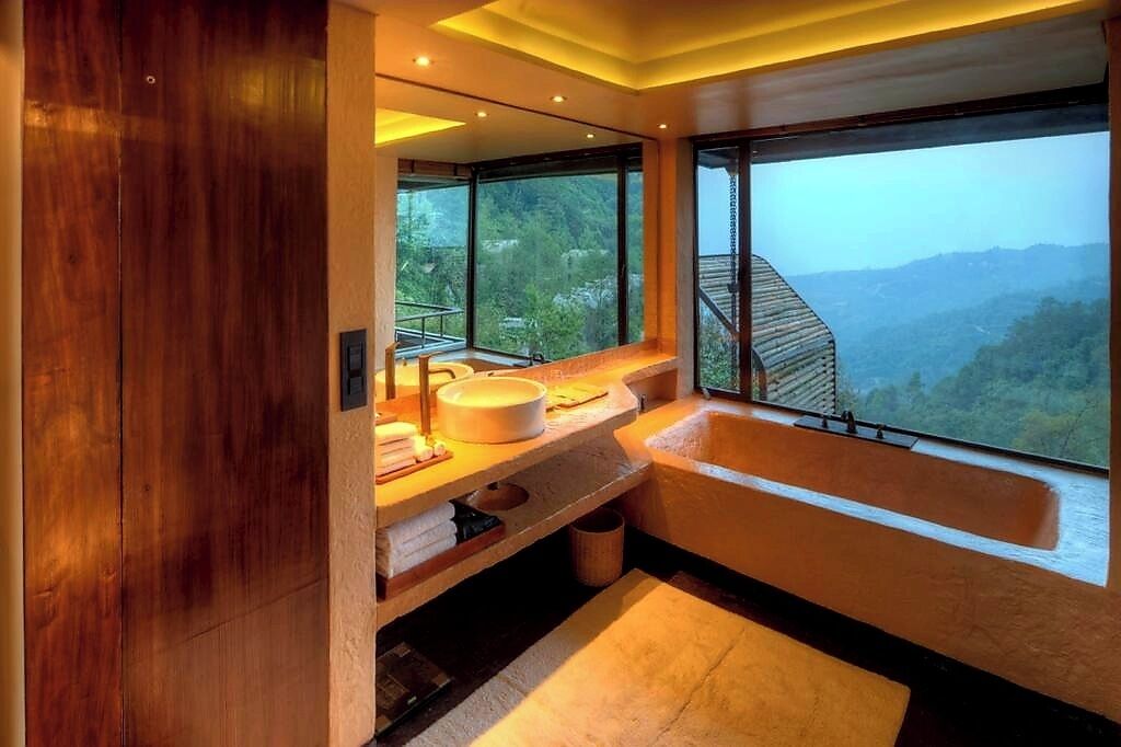 A modern bathroom with a large bathtub, a sink, and a mirror. It features a large window with a view of a mountainous landscape.