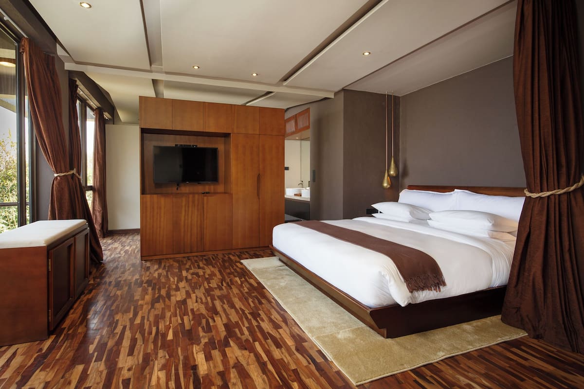 A bedroom with a large bed, wooden flooring, a TV mounted on a wooden panel, and a view of the outdoors through large windows.