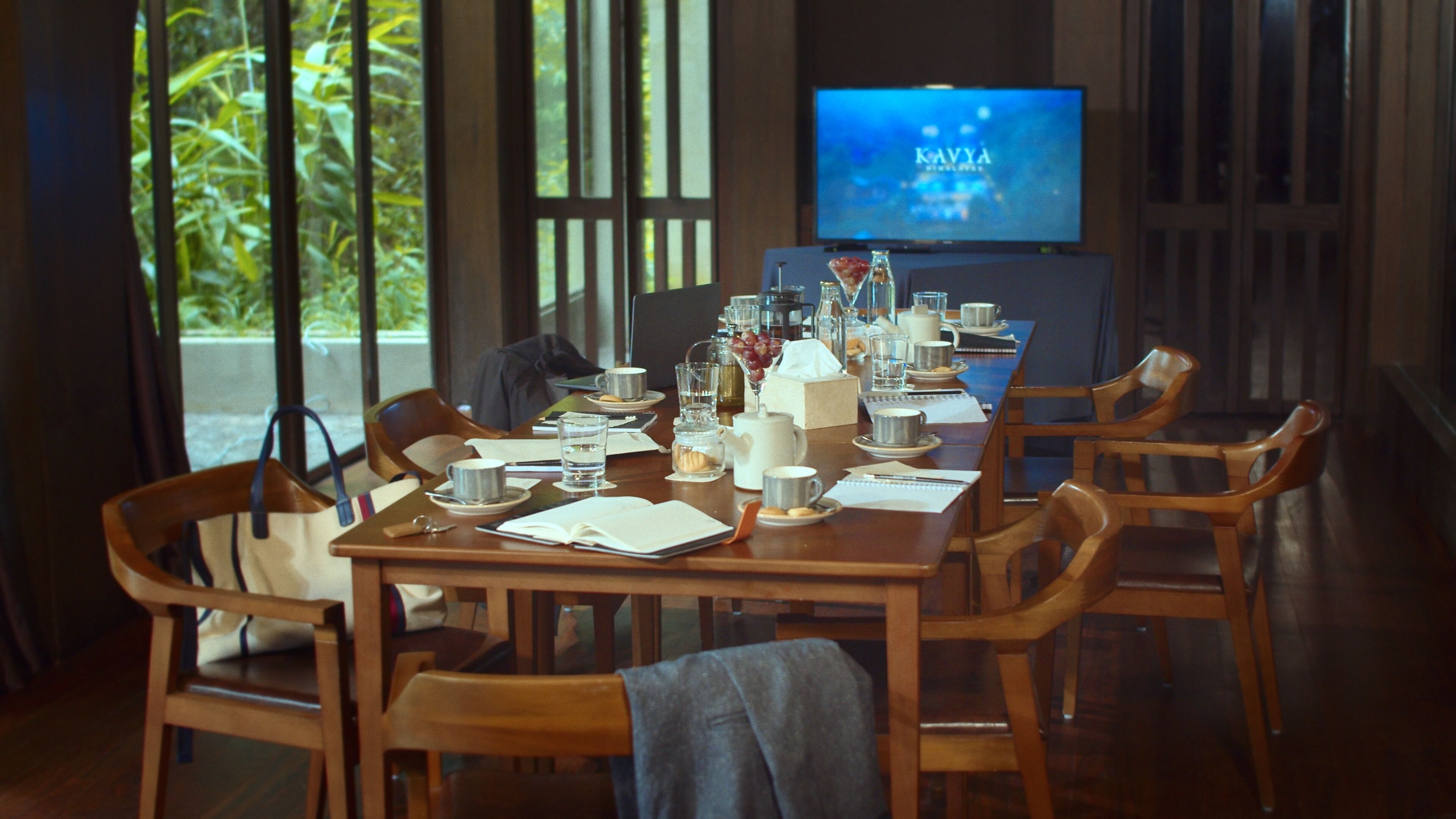 Un coin repas avec une longue table dressée pour un repas, des chaises disposées autour, et un téléviseur qui affiche le mot « KAVYA » en arrière-plan.