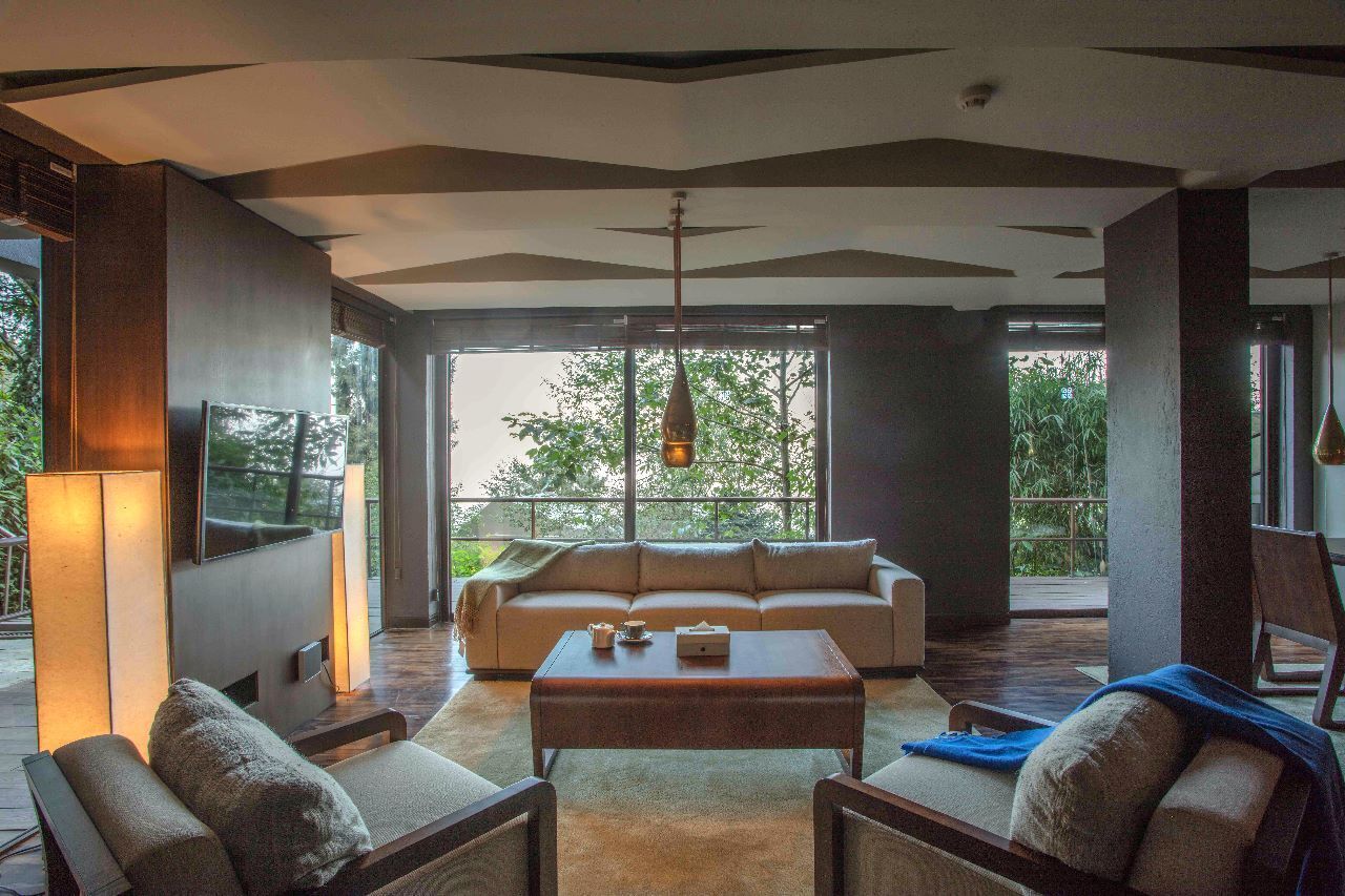 Una sala de estar moderna con un sofá, una mesa de centro y sillones, ventanas amplias con vistas a zonas verdes y un televisor de pantalla plana.