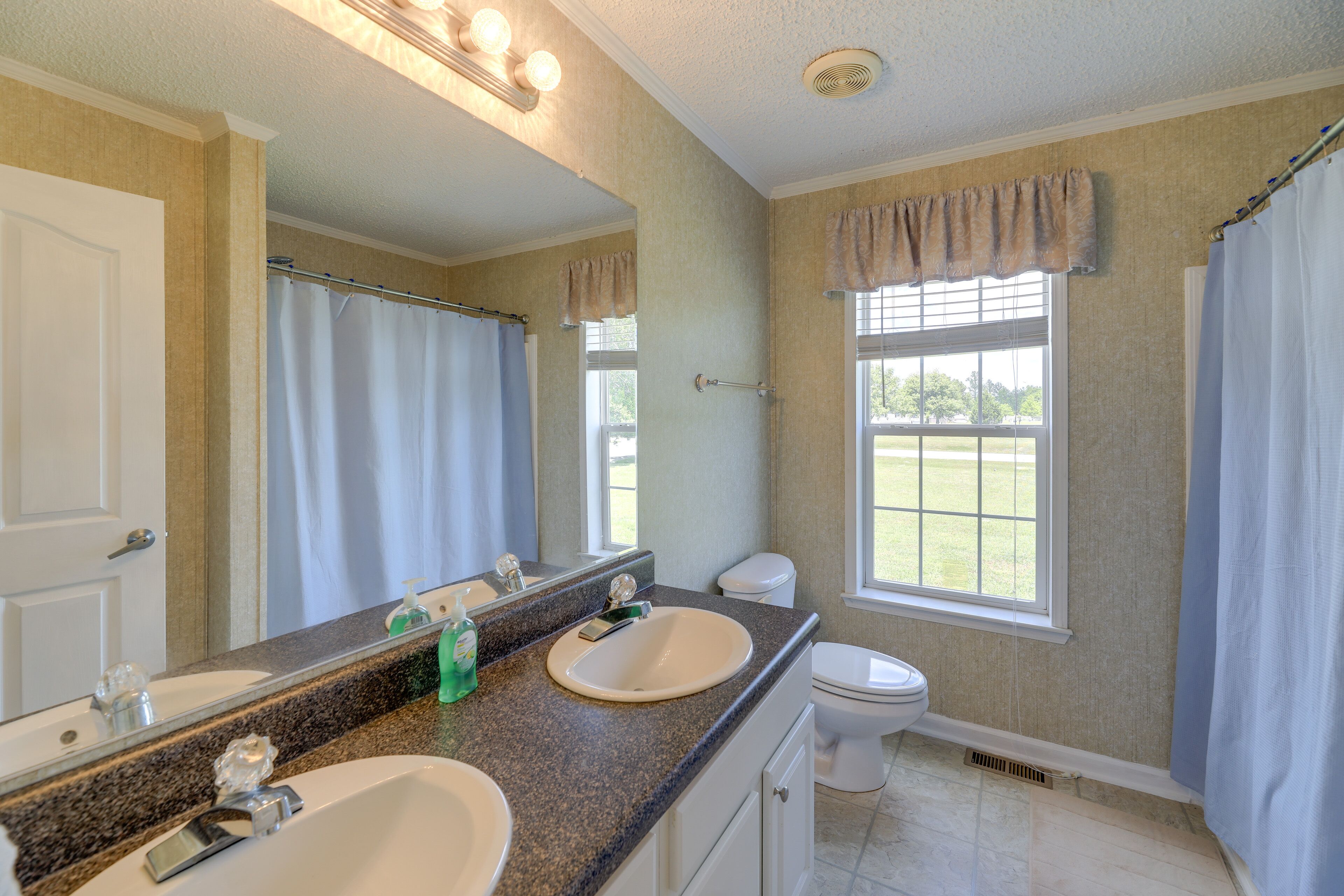 A bathroom with two sinks, a toilet, a large mirror, and a window with curtains.