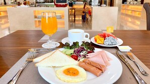 A breakfast setting with toast, eggs, ham, a salad, and fruit on a plate, a glass of orange juice, and a cup of coffee on a table in a hotel lobby. - ST Vegas Hotel (Hadxayfong)
