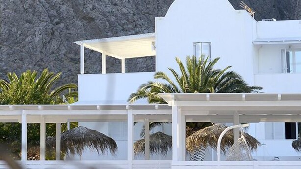 A modern white building with a flat roof and a balcony, surrounded by palm trees and a rocky cliff. - Aspro Phos (Santorini)