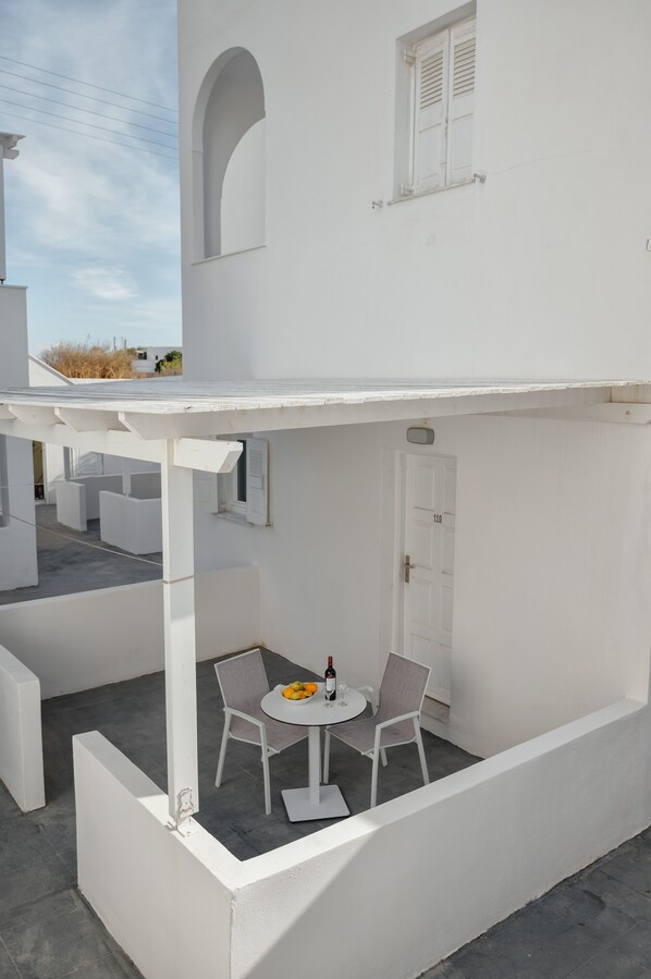 A white building with a covered patio area featuring a table and chairs, a door, and a window with shutters.