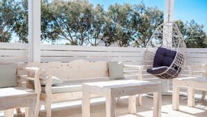 An outdoor seating area with wooden furniture, including benches and a hanging chair, under a white structure with a view of trees. - Aspro Phos (Santorini)