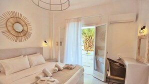 A hotel room with a bed, a wall-mounted mirror, a door leading to a balcony, and a small table with a chair. - Aspro Phos (Santorini)