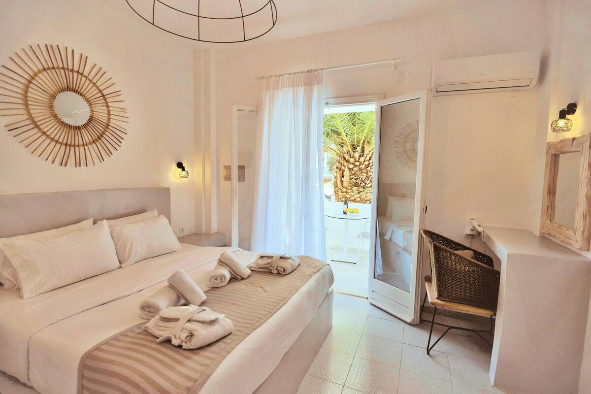 A hotel room with a bed, a wall-mounted mirror, a door leading to a balcony, and a small table with a chair.