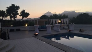 Zona de piscina en la azotea con vistas a montañas, muebles de exterior blancos y una pequeña chimenea.