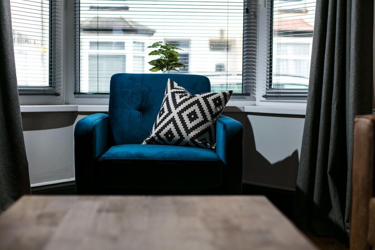 A blue armchair with a black and white patterned pillow, positioned by a window with blinds, and a small potted plant on the windowsill.