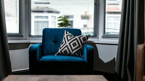 A blue armchair with a black and white patterned pillow, positioned by a window with blinds, and a small potted plant on the windowsill. - Sutton House by Pureserviced (Plymouth)