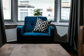 A blue armchair with a black and white patterned pillow, positioned by a window with blinds, and a small potted plant on the windowsill. - Sutton House by Pureserviced (Plymouth)