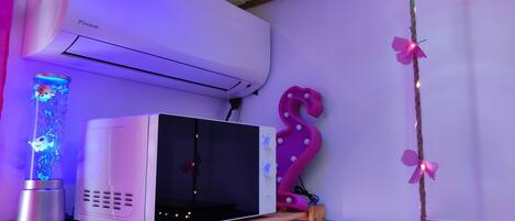 A room with a white air conditioner, a microwave on a wooden shelf, a pink decorative item, and a string of lights.