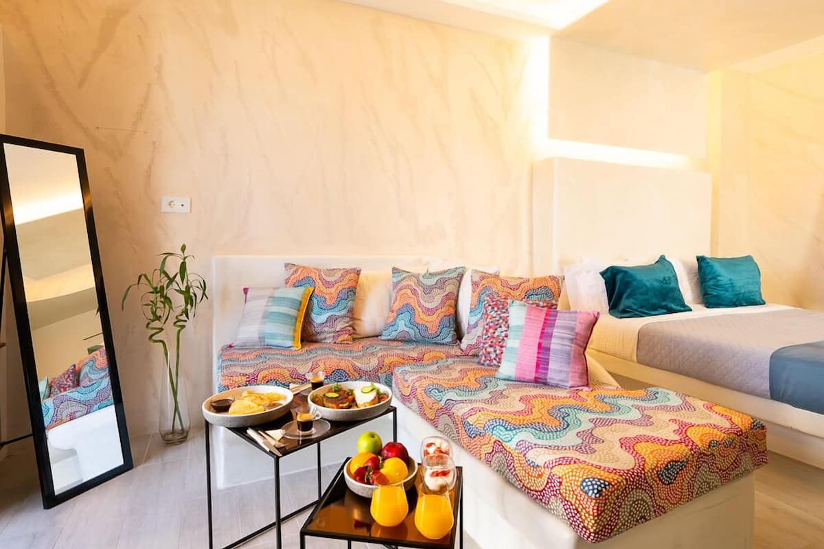Una habitación de hotel moderna con un conjunto de sofás, una mesa de comedor con frutas y bebidas, y un espejo.