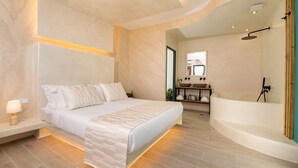 Un dormitorio moderno con una cama grande, un banco empotrado, una mesita de noche con lámpara y vistas a un baño con bañera y ducha.