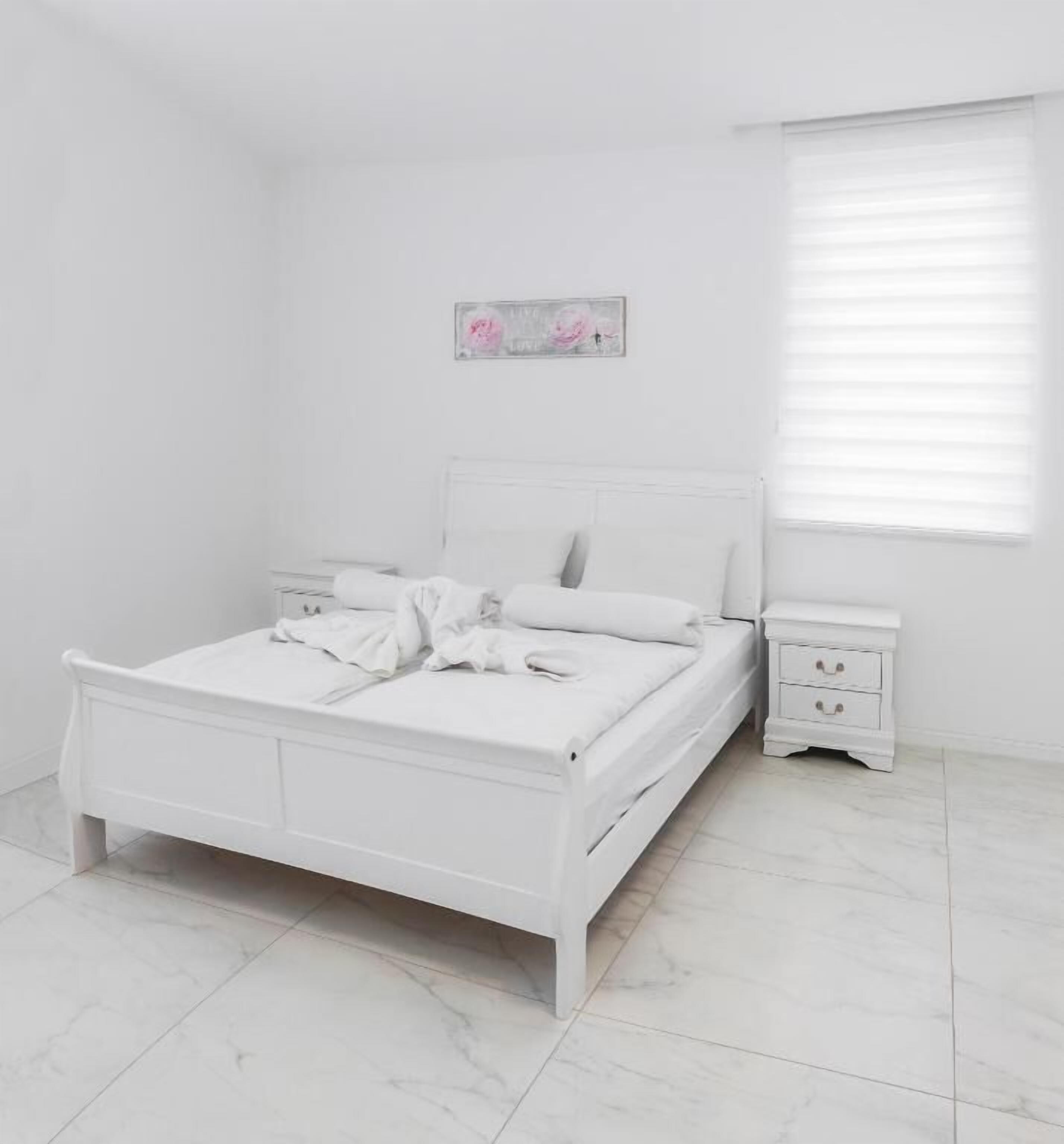 Un dormitorio blanco con cama, mesita de noche y ventana con persianas.