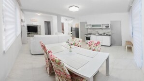 A modern, well-lit living room with a white sofa, a dining table with floral chairs, and a kitchen area with white cabinets and appliances.