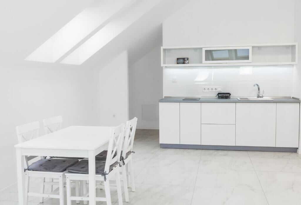 Une cuisine moderne avec des meubles blancs, une table à manger blanche et des chaises. On y trouve un petit plan de travail avec un évier et une bouilloire.