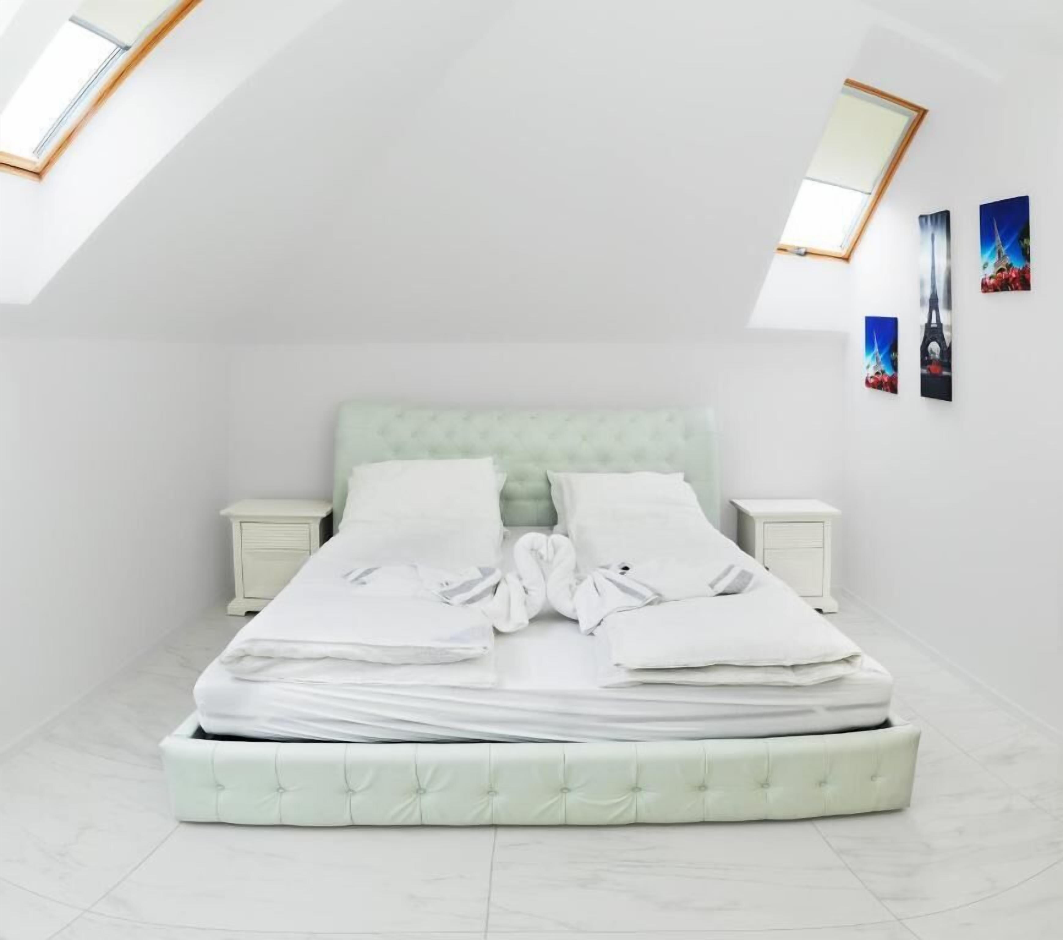 Cama bem arrumada com lençóis e almofadas brancas, cabeceira estofada em verde claro e duas mesas de cabeceira com mesinhas de apoio brancas.