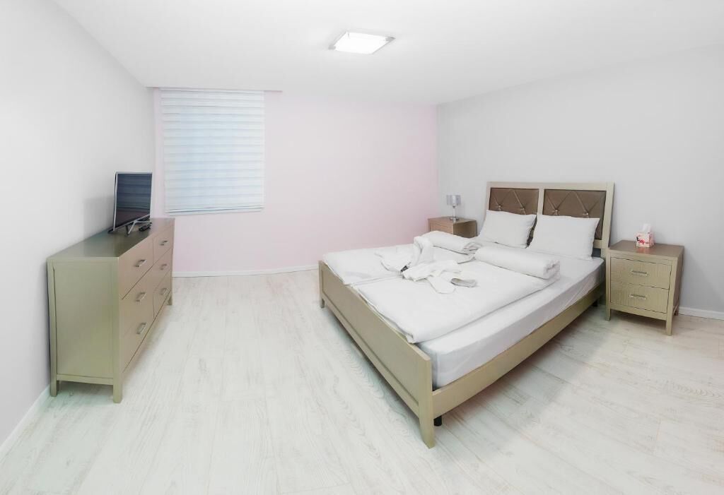 A bedroom with a bed, bedside tables, a dresser, a TV, and a window with blinds.