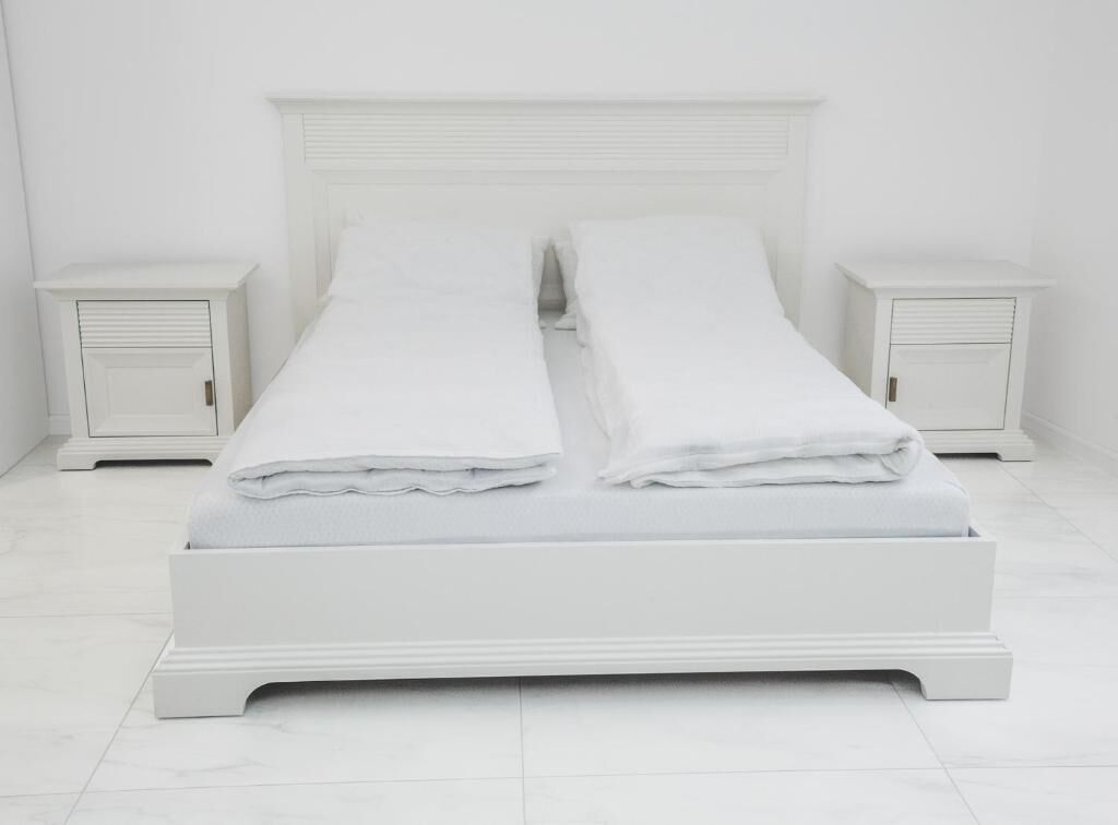 Una cama blanca con dos almohadas y ropa de cama blanca doblada, flanqueada por dos mesitas de noche con cajones, todo sobre un fondo de pared y suelo blancos.