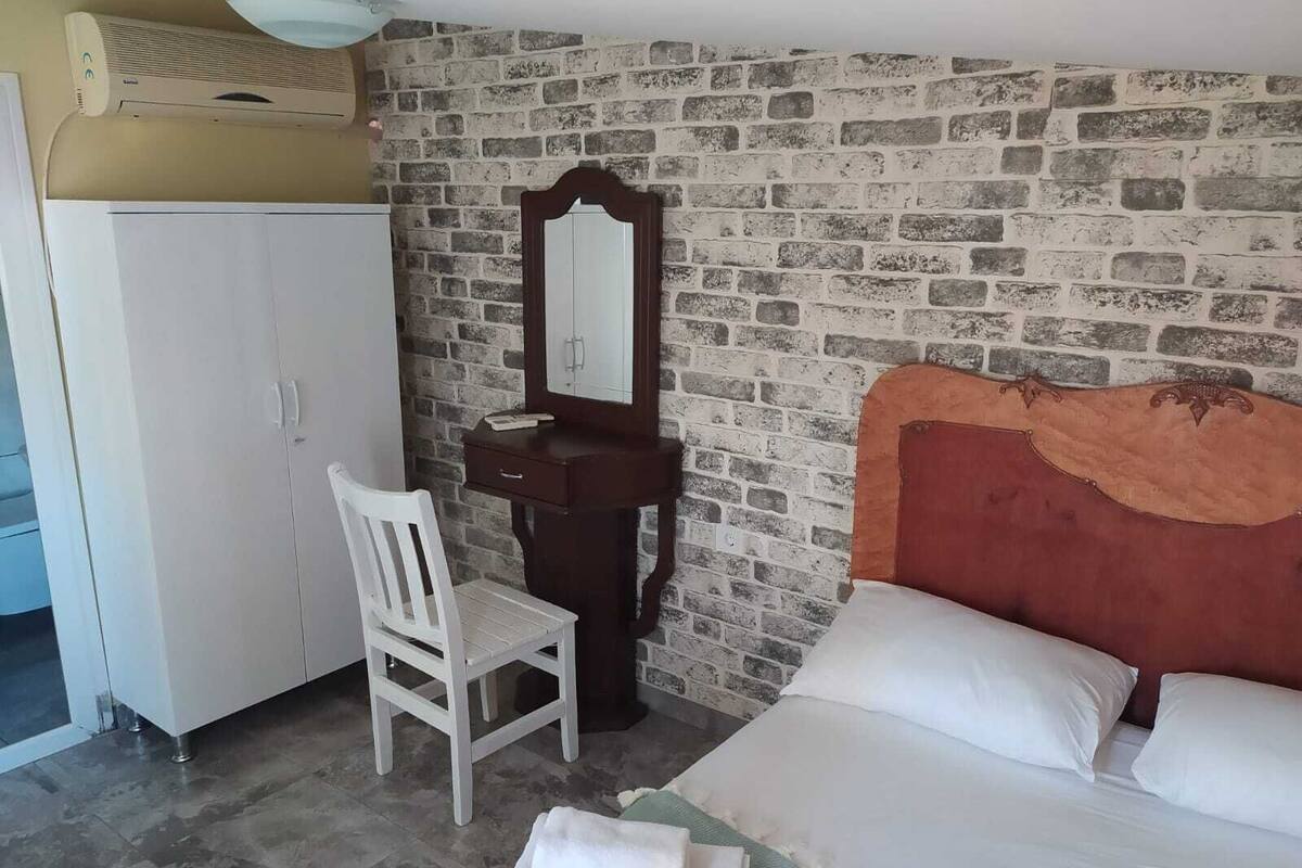 A room with a brick-patterned wall, a white wardrobe, a wooden dresser with a mirror, a white chair, and a bed with white bedding.