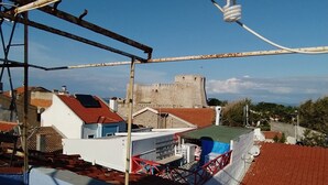 Un toit-terrasse surmonté d’un toit de tuiles rouges, un bâtiment blanc avec une porte bleue, et, au loin, un grand château en pierre.
