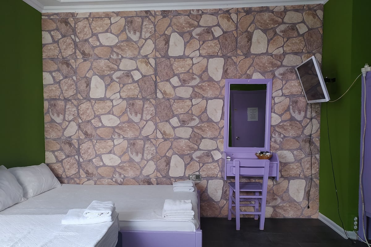 A room with a stone wall, a bed with white linens, a purple vanity with a mirror, and a small table with toiletries.