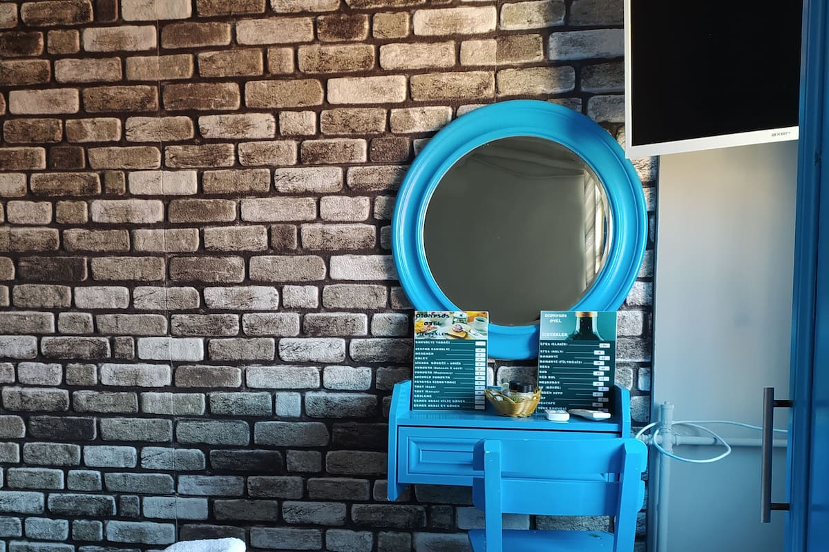A room with a brick wall, a blue vanity with a mirror, a bed with white linens, and a small table with a basket.