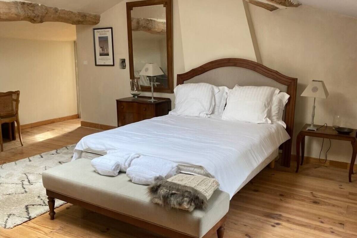 Un dormitorio con cama de madera, ropa de cama blanca, una alfombra pequeña, una mesita de noche de madera, una silla y un espejo en la pared.