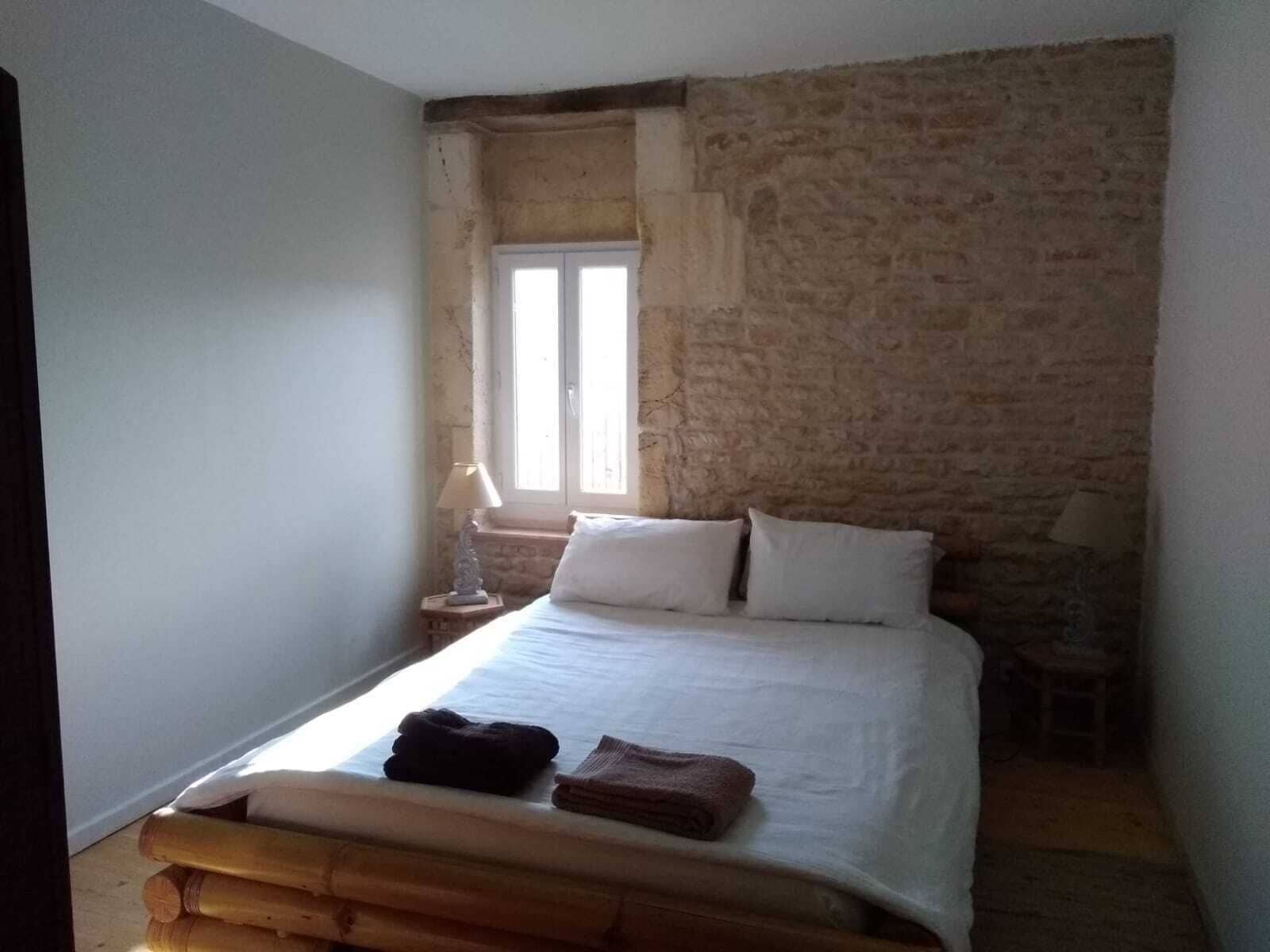 A bedroom with a stone wall, a bed with white linens, two pillows, a nightstand with a lamp, and a window with curtains.