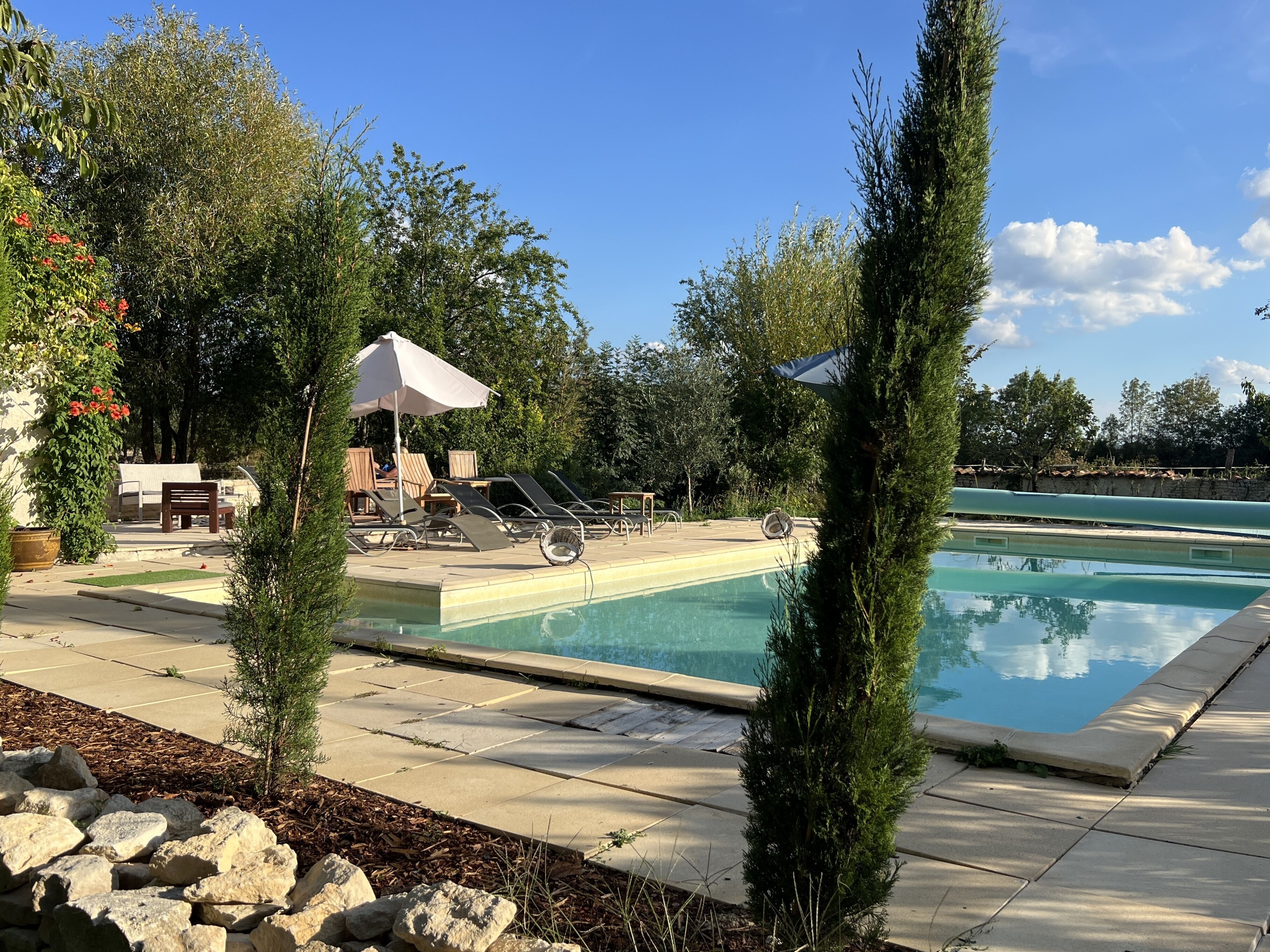 A swimming pool with lounge chairs and umbrellas, surrounded by trees and a stone wall.
