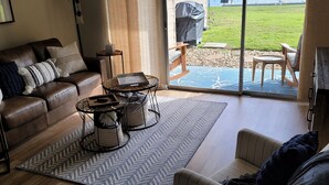 A spacious living room with a brown leather couch, a striped armchair, and a coffee table with woven baskets. A large sliding glass door leads to a patio with a grill and outdoor seating. - Ground Floor Waterfront Condo! (Montgomery)