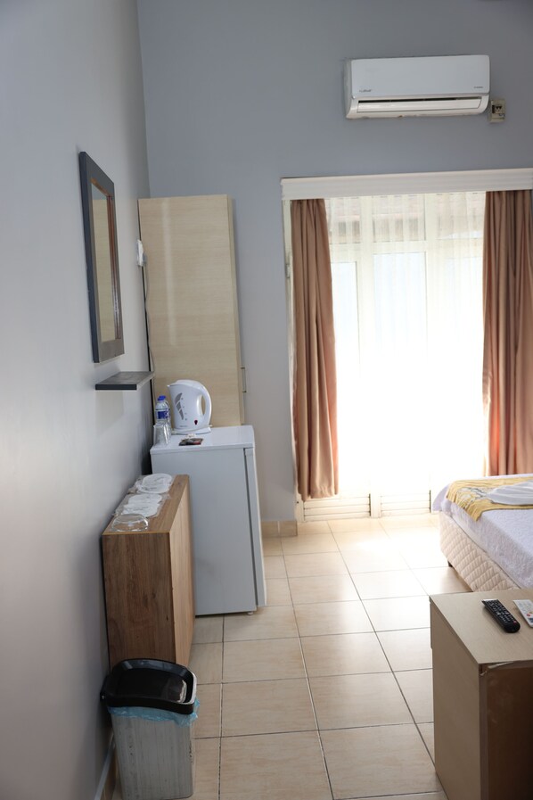 A hotel room with a bed, a small desk with a kettle, a trash bin, and a window with curtains.
