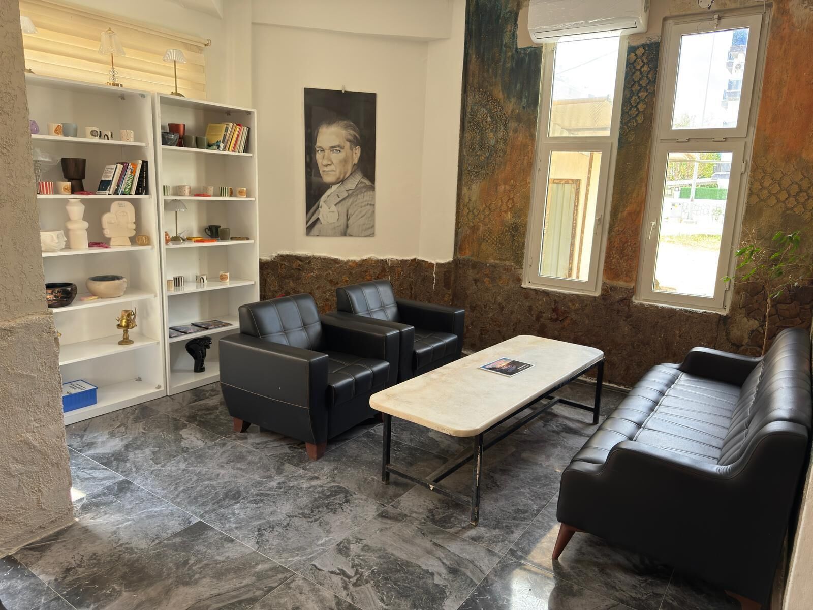 A room with a stone wall, a black leather sofa, a black leather armchair, a white coffee table, and a bookshelf with various items.