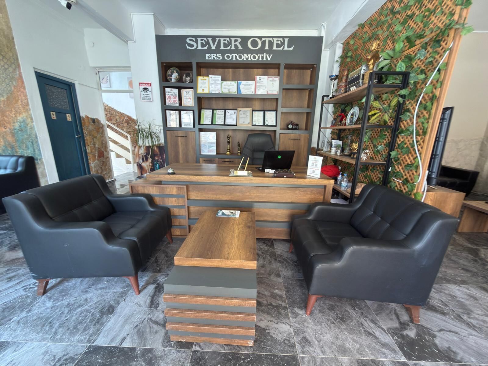 A hotel reception area with a desk, two black leather sofas, a coffee table, and a wall with certificates and awards.